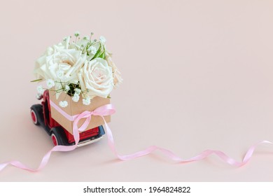 Red Car With A Gift Box Of Roses Flowers On The Roof On Pink Background. Happy Valentine's Day, Mother's Day, March 8, World Women's Day Holiday Card Concept, Flower Delivery.