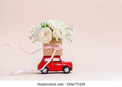 Red Car With A Gift Box Of Roses Flowers On The Roof On Pink Background. Happy Valentine's Day, Mother's Day, March 8, World Women's Day Holiday Card Concept, Flower Delivery.