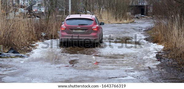 A red\
car runs over a puddle on a muddy rural road, Dalnevostochniy\
prospekt, Saint Petersburg, Russia, February\
2020