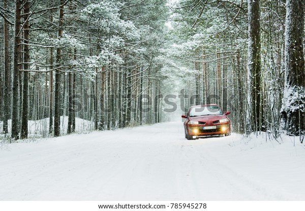 Red car with yellow headlights on snow-covered\
forest road.