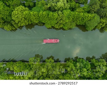 Red cargo barge navigating through turquoise waters surrounded by lush green forest along Hangzhou's Shangtang River, captured from above.
