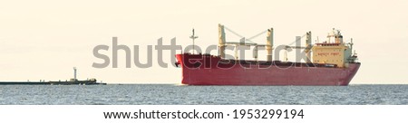 Red cargo ship (bulk carrier) sailing in the Baltic sea. Lighthouse in the background. Freight transportation, nautical vessel, logistics, industry, commerce, environmental damage. Panoramic view