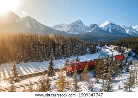 Red cargo train passing through Morant's curve in Bow Valley, Banff National Park, Alberta, Canada. Rocky Mountains of North America. Beautiful snowy winter scenery in background with lens reflection.
