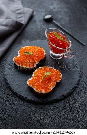 Red caviar in a bowl and caviar sandwiches on a black stone board. On a black structural background