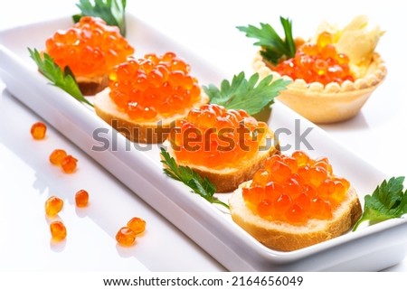 Red caviar on butter buns decorated with green seasonings on white rectangular plate. In background is tartlet with butter and red caviar decorated with parsley on side on white background.