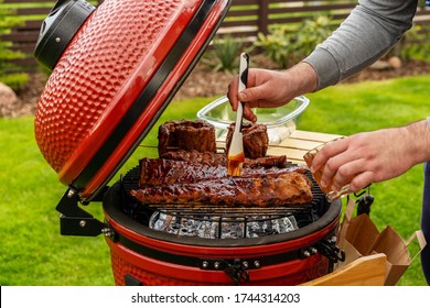 Red Ceramic Barbecue Grill. The man coats pork ribs with BBQ sauce. Grilling, Smoking, Baking, BBQ and Roasting process. Post-quarantine Picnic in modern homes terrace. Lifestyle concept. - Shutterstock ID 1744314203