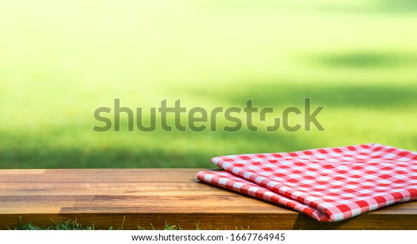 Red checked tablecloth on\
wood with blur green courtyard background.Summer and picnic\
concepts.Design for key visual food and drink products.no\
people