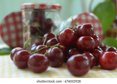  red cherries with stems and leaf on yellow white dicey tablecloth jars with cherries, red white caps,basket, and green background