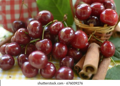 red cherries with stems and leaf on yellow white dicey tablecloth jars with cherries,basket with cherries, and red white caps,cinnamon rods