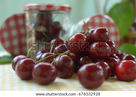  red cherries with stems and leaf on yellow white dicey tablecloth jars with cherries, red white caps,basket, and green background