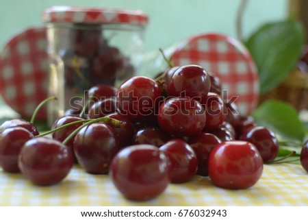 red cherries with stems and leaf on yellow white dicey tablecloth jars with cherries,basket, and red white caps and green background