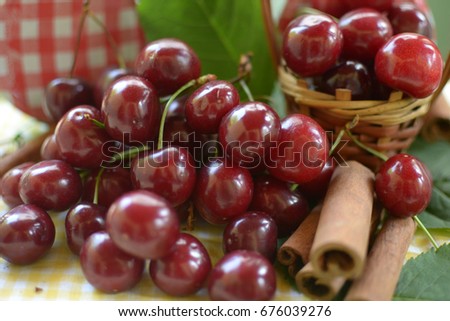 red cherries with stems and leaf on yellow white dicey tablecloth jars with cherries,basket with cherries, and red white caps,cinnamon rods