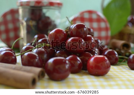 red cherries with stems and leaf on yellow white dicey tablecloth jars with cherries,basket, and red white caps,cinnamon rolls and green background
