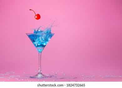 Red Cherry Falling Into A Blue Coktail Splashing In Martini Cup Isolated On Wet Pink Background With Space For Text. Concept Image For Entertainment Club And Party.