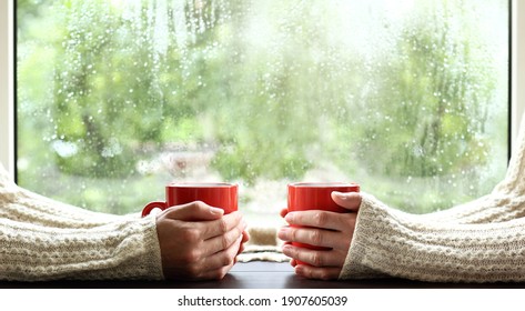 Red Circles In The Hands Of Those Dressed In Sweaters Against The Background Of A Window With Drops After The Rain. A Warming Drink For A Cozy Conversation