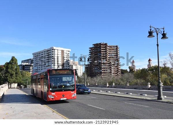 Red city bus travels down a street in city of\
Valencia. Public transport and city buses in Eurupe. Сity street,\
road traffic, cars on road, people and buildings. Dec 09, 2021,\
Spain, Valencia.\
