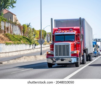 Red Classic Industrial Big Rig Semi Truck Tractor Transporting Cargo In Dry Van Semi Trailer Running On The Divided Highway Road In Front Of Another Semi Trucks Going In The Convoy Line