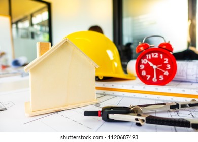 Red clock on archetecture blueprint with pencil, yellow helmet, wooden house model and engineering equipments in modern working office room  - Powered by Shutterstock