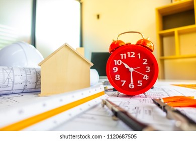 Red clock on archetecture blueprint with pencil, yellow helmet, wooden house model and engineering equipments in modern working office room  - Powered by Shutterstock