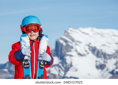 In a red coat, a child smiling, gripping skis with majestic, snow-covered mountains in the background winter portrait - Powered by Shutterstock