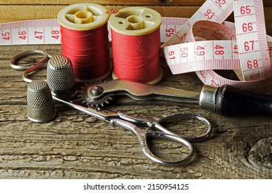 RED COLORED THREAD ON REELS WITH RED AND WHITE MEASURINGTAPE, THIMBLES, A TRACING WHEEL AND SCISSORS ON A WOODEN BOARD
