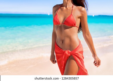 Red coral bikini body sexy swimsuit model woman showing off toned stomach abs and slim waist wearing sarong pareo fashion clothes on beach. Lower body crop of torso and hips.