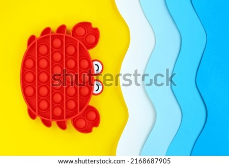 Red crab popular antistress sensory pop it toy on seashore made of color paper cut, blue waves and yellow sand. For children and adults relaxation and fun on vacation. Summer holidays concept.