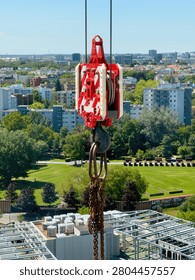 Red crane hook block and chain hanging high above a green city park and residential buildings on a construction site in summer