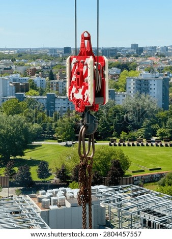 Red crane hook block and chain hanging high above a green city park and residential buildings on a construction site in summer