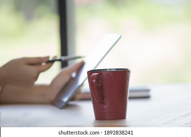 A Red Cup On Table With Woman Hand Holding Pen Pointing To Tablet With Coffee Cup In Cafe As Background. Work From Hom Idea.