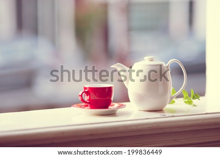 Red cup of tea with white teapot on wooden windowsill with blurred city in a background.  Vintage look.
