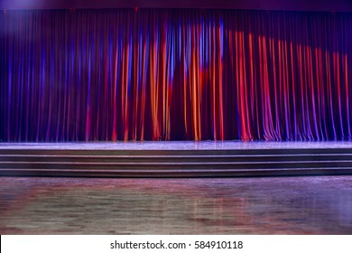 Red Curtains And The Stage Parquet With Stairs In Theater With Colorful Lighting.