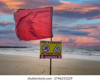 Red danger flag and Rip Current — No Swim Zone warning sign on North Goa beach at dramatic sunset. 
