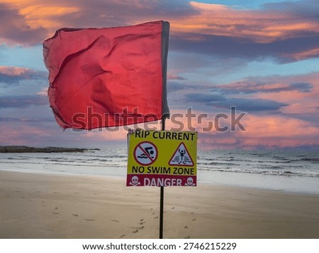 Red danger flag and Rip Current — No Swim Zone warning sign on North Goa beach at dramatic sunset. 