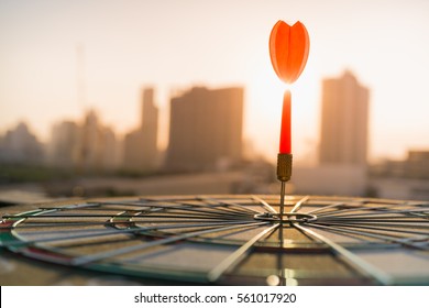 Red dart arrow hitting in the target center of dartboard with modern city and sunset background. Target business, achieve and victory concept .