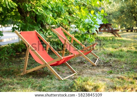 Red Deckchairs in autumn garden. Two deckchairs on summer green lawn. Lounge sunbed. Wooden garden furniture on grass lawn outdoor for relax. Backyard exterior. Garden landscape with chairs in nature.