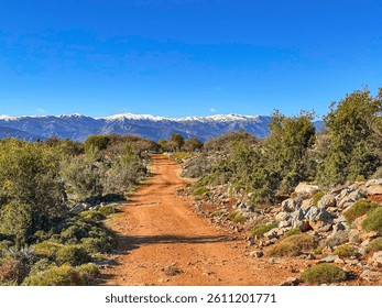 Red dirt path winds through rocky countryside leading towards snowy mountains. Offroad trail shows a stunning contrast between dry Mediterranean scrub and remote Alpine landscape under blue skies.