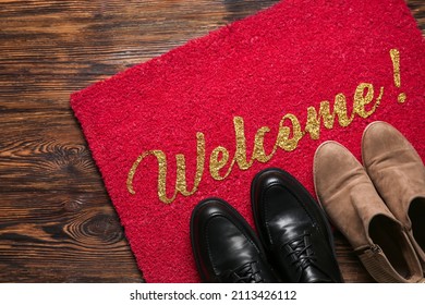 Red Door Mat With Word WELCOME And Shoes On Dark Wooden Floor