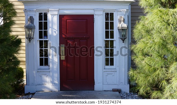 A red door with white trim adorned by\
lanterns on either side, and green\
shrubs.