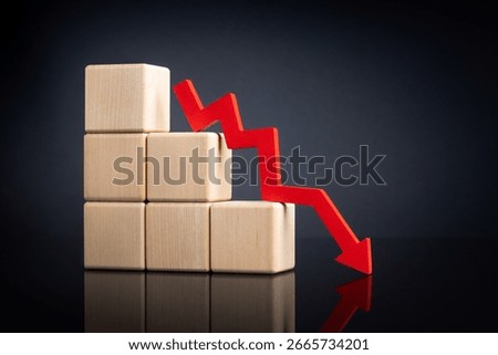 A red downward zigzag arrow next to with stacked wooden blocks, symbolizing a declining trend on a reflective dark surface. High quality photo