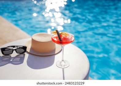 A red drink in an elegant glass with a long stem on the side of the pool. An alcoholic ice drink decorated with a flower on a background of turquoise water. Magic bokeh and straw hat. - Powered by Shutterstock