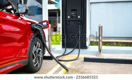 A  red electric car is charging at a charging station.Electric vehicle recharge battery at a public charging station in the city area. EV car attached with electric charger for eco-friendly idea.
