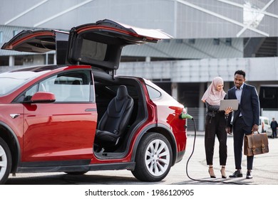 Red electric car is charging while muslim woman and african man talking near. Female seller in hijab using laptop while demonstrating modern auto to male customer in suit. - Powered by Shutterstock