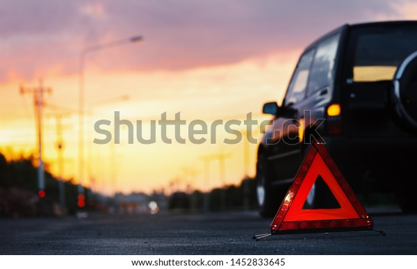 Red emergency stop sign (red\
triangle warning sign) and broken black SUV car  on country\
road.