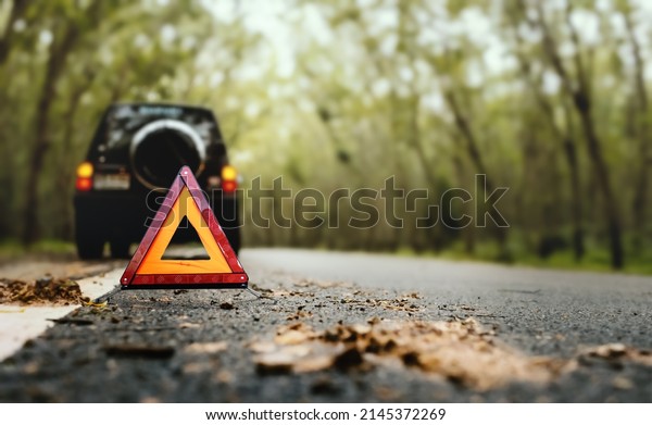 Red emergency stop sign (red\
triangle warning sign) and broken black SUV car  on country\
road.
