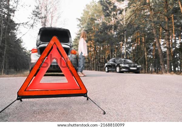 Red\
emergency triangle on the road amid a blurry car and a woman in\
despair catching passing cars waiting for help. Focus on the\
emergency sign. Accident and broken car on the\
road.