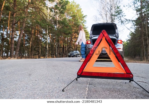 Red\
emergency triangle on the road amid a blurry car and a woman in\
despair catching passing cars waiting for help. Focus on the\
emergency sign. Accident and broken car on the\
road.