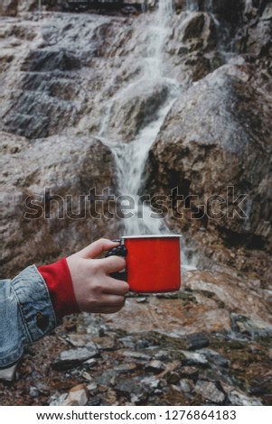 Red enameled camping mug on background of  waterfall