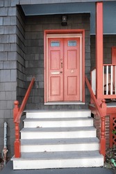 Red Entrance Door With A Round Window Of A House With A Wooden Facade. With Stairs And Railings At The Entrance.