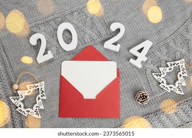 A red envelope with an empty sheet of paper for congratulations on Christmas and New Year. Wooden numbers of the new year 2024 on a gray knitted background, bokeh lights. - Powered by Shutterstock